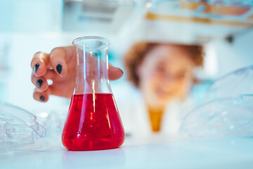 Young female scientist working at the laboratory. Close-up of serious laboratory worker holding ampoule in front of eyes and examines contents. Scientist in protection gloves eyewear and white coat. 