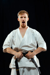 Portrait of young man, karate fighter in white kimono and black belt in fighting stance on dark background. Sport, action, combat sports, ad concept © master1305