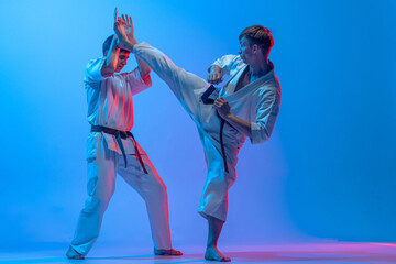 Fototapeta premium Studio shot of sports training of two karatedo fighters in doboks isolated on blue background in neon. Concept of combat sport, challenges, skills