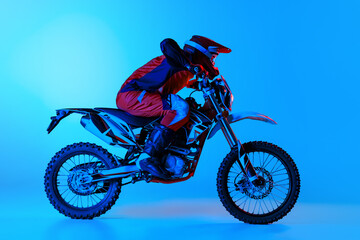 Portrait of young man, biker in full equipment riding motorbike isolated over blue studio background in neon light. Motocross