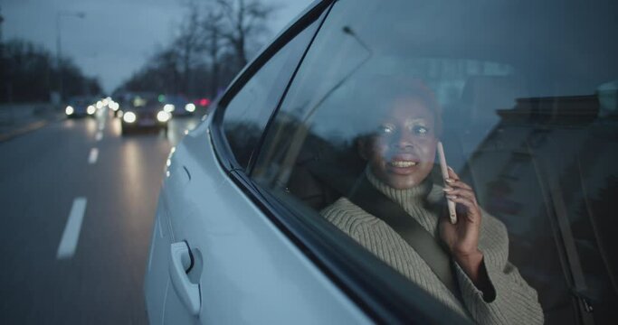 Positive young black woman is having a call on her phone while beeing a passenger in a taxi