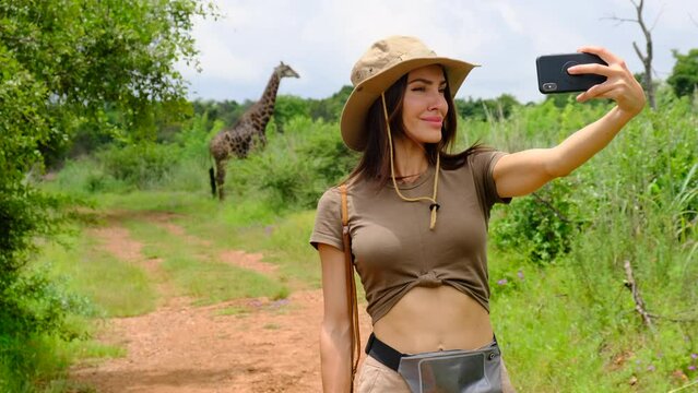 Happy zoology student girl taking selfie photo on smartphone while giraffe. a woman traveler in safari clothes and a hat makes a photo on the phone against the backdrop of a tall giraffe in the jungle