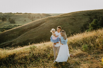 A young family with a little daughter are hugging and walking in the field at sunset.