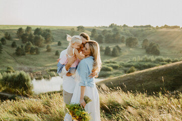A young family with a little daughter are hugging and walking in the field at sunset.