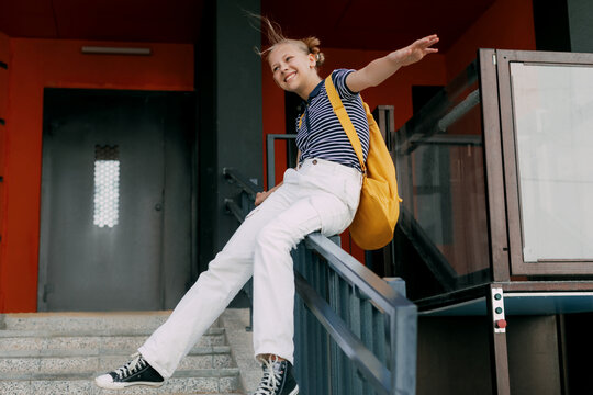 A teenage girl laughs and slides down the railing on her way to school. A cheerful girl hurries to school
