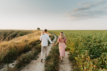 A young family with a little daughter are hugging and walking in the field at sunset.