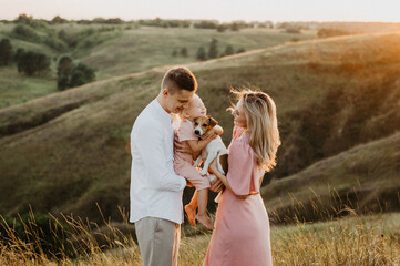 A young family with a little daughter and a dog are hugging and walking in the field at sunset.