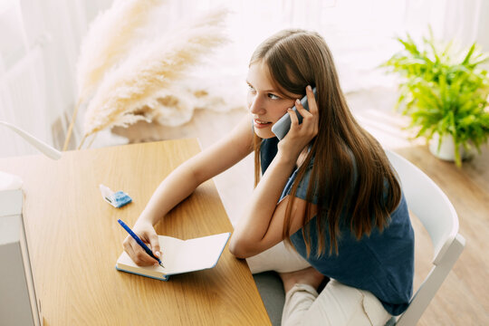 A young attractive student talks on a smartphone while studying at home, discusses homework or a joint project with friends, makes notes in a notebook