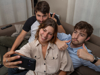 trois jeunes amis font un selfie assis dans un sofa. Moment fun de complicité entre jeunes adultes - obrazy, fototapety, plakaty