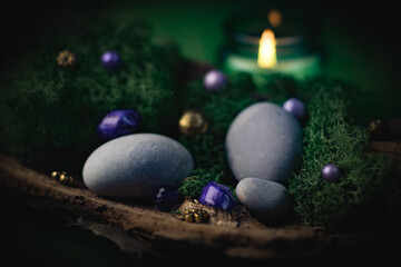Lilac, golden stone lies in the bark of a tree with green moss, gray stones and a burning candle on a green