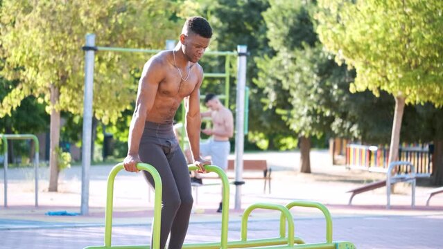 Young fit shirtless black man doing calisthenics workout on parallel bars outdoors on sunny day. Fitness and sport lifestyle.