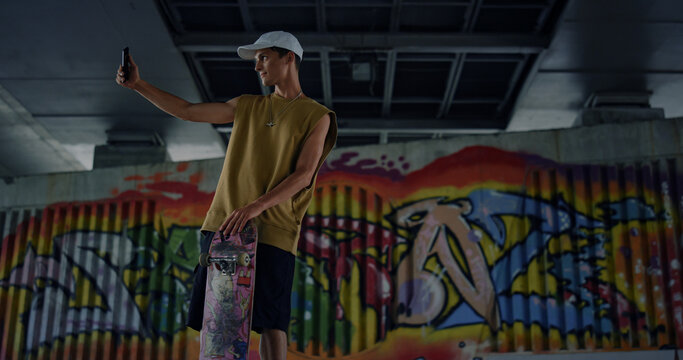 Casual skater taking selfie photo on smartphone at skate park graffiti wall. 