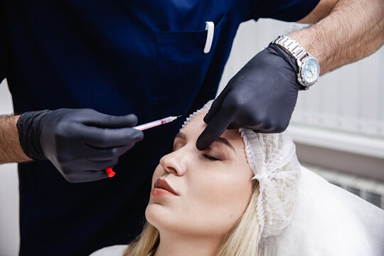 A cosmetologist in black gloves injects botulinum toxin into the patient's forehead. A blonde patient in a disposable cap lies with her eyes closed