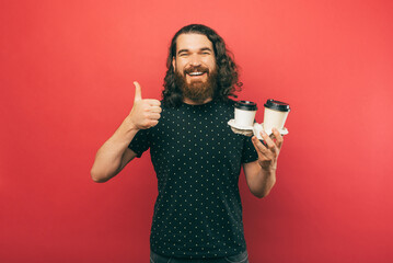 A photo of a bearded young man holdng some cups of coffee while showing a thumb up and smiling
