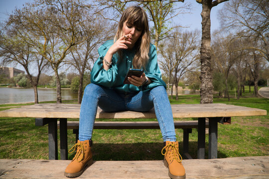 A young, pretty, blonde woman is sitting at a wooden table in the park, looking at her mobile phone and smoking a cigarette. In the background you can see the river on a sunny day. Stop smoking