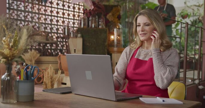 Owner of small business taking online orders in front of laptop computer. Female entrepreneur of local flower shop talking with customer or partner online wearing apron talking on phone