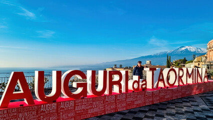 Man with christmas tree and signs of Christmas wishes (Auguri) on the main square (Piazza IX Aprile) in Taormina, Province of Messina, Sicily, Italy, Europe, EU. Holidays at the Mediterranean sea