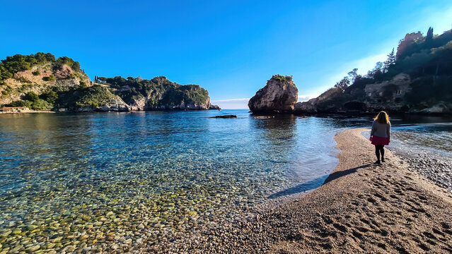 Woman in dress walking on stone pebble beach Spiaggia di Isola Bella towards the paradise island Isola Bella in Taormina, Sicily, Italy, Europe, EU. Dreamy seascape at Ionian Mediterranean sea