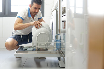 Kneeling man placing a dish in the automatic dishwasher in his kitchen. Hand of a person holding a...