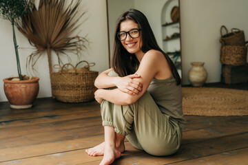 A young twenty year old brunette girl in glasses sits cross-legged on the floor and smiles.