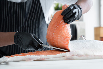 Selective focus on chef man hand with black glove holding fresh salmon while another hand holding knife skillfully to cut salmon at kitchen. Anonymous chef prepare raw ingredients for japanese food