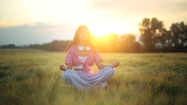 Young healthy beautiful girl having a yoga meditation session in the field during sunrise - healthy lifestyle, zen concept. Yoga meditation practice in nature. Woman Enjoying Summer Sunset View