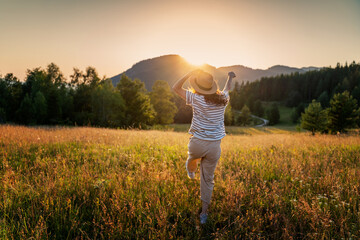 Happy young woman in a hat enjoying a summer evening and sunset in the mountains on a field