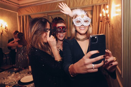 Positive women in carnival masks taking selfie in restaurant - Powered by Adobe