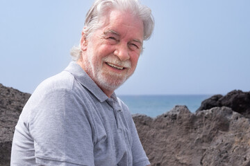 Fototapeta na wymiar Portrait of bearded white-haired caucasian man sitting on the rocks along the sea looking at camera. Smiling retiree enjoying freedom and good time