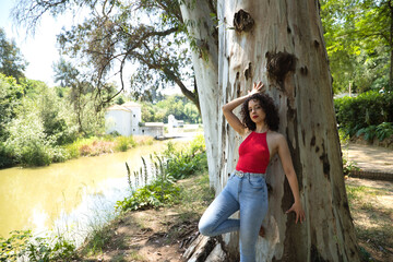 Young, attractive, brunette woman with curly hair, red t-shirt and jeans, leaning against a big tree in an outdoor park with provocative look. Concept sensuality, provocation, beauty, nature.