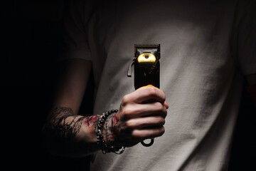 hand of young barber man with stylish tattoo in white t shirt holding electric hair clipper, razor for hairdressers on dark background. Barbershop beauty salon. The barber clippers instead of scissors - obrazy, fototapety, plakaty