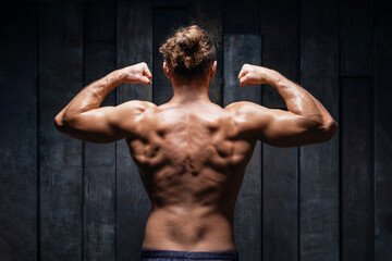 Fototapeta na wymiar Sexy muscular bodybuilder posing from back on dark background. Handsome male with perfect body after training. Athlete do exercise fitness. Male training upper body working out at hard cross-fit.