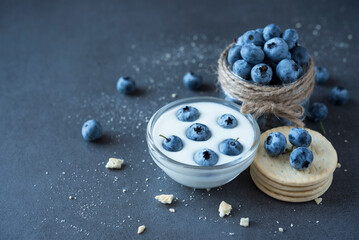 Blueberry in a glass jar with yogurt, another one jar with blueberry, cookies on the dark grey...