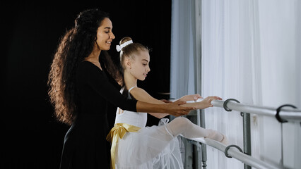 Teen girl child teenager ballerina in tutu has advice of woman coach teacher learn to dance in dancing school near ballet barre stretching leg hip. Female trainer helps with stretch correct position