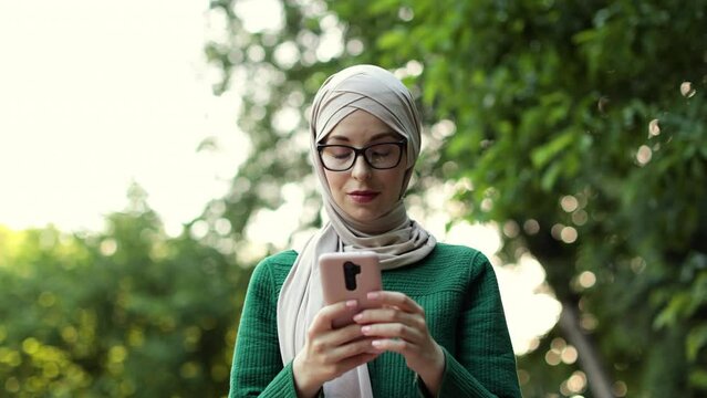 Attractive smiling muslim woman in hijab texting on smart phone standing on background of green city park. Portrait of young arabian businesswoman using mobile phone outdoor.