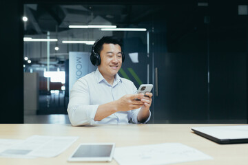 A break at work. Young handsome Asian man, freelancer listening to music on the phone in headphones. Dances along, smiles. Sitting at a desk in a modern office