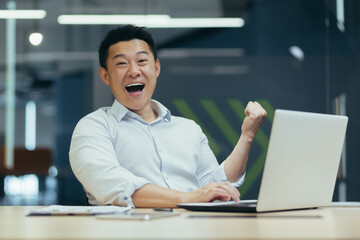 Successful and happy young Asian man, businessman, rejoices, shows yes with his hand, looks at the camera. Made a contract. Won money. Sitting with a notebook at the table in a modern office.