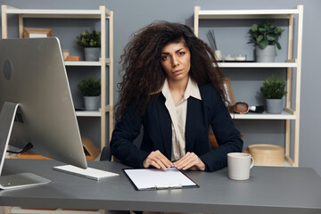 Serious angry irritated tanned adorable curly Latin businesswoman in jacket looks at camera in office interior. Copy space Mockup Banner. Corporation leader lady uses modern computer in work