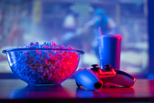 Joystick, Gamepad, Popcorn In A Large Glass Bowl, Soda Drink In A Blue Plastic Glass Against The Background Of A Plasma TV Screen. Neon Lighting. Recreation, Video Games With Fast Food.