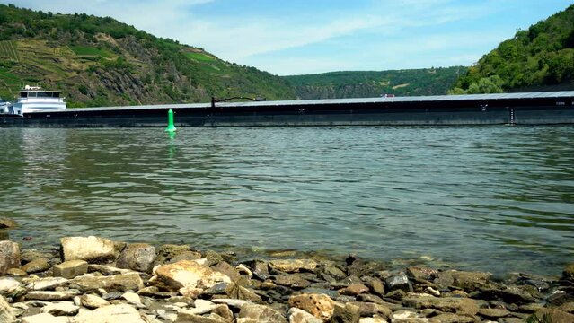 A freight ship or barge passing close by a buoy in low water levels on Rhine during drought in summer 2022 near Oberwesel and Loreley revealing a sandbank behind it and vineyards on the mountainside
