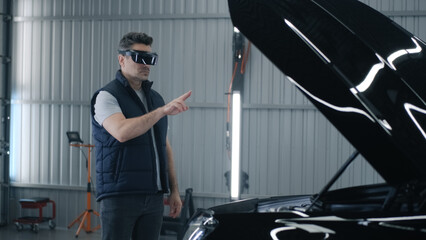A male mechanic in a virtual reality headset gesturing and standing near the open hood of a car,...