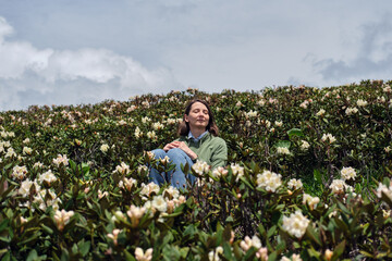 Happy middle-aged woman sitting with her eyes closed among blooming wild rhododendrons. Mountain hiking, outdoor activities, mental health concept.