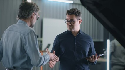 Satisfied service manager in repair shop and client shaking hands after diagnosing and fixing a car in a car service, and manager with a tablet looking at the camera