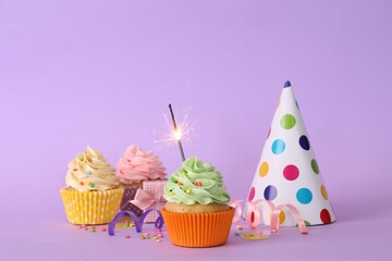 Birthday cupcakes with burning sparkler, party hat and gift box on violet background