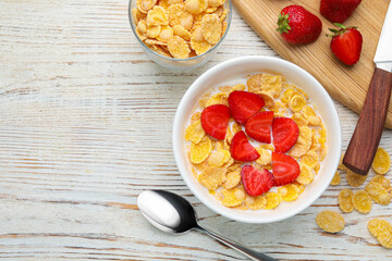Bowl of tasty crispy corn flakes with milk and strawberries on white wooden table, flat lay. Space...