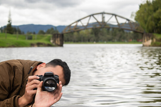 Joven fotógrafo tomando fotos con una cámara profesional  en el lago de un parque natural con un puente de fondo - Parque Simón Bolivar, Bogotá, Colombia 