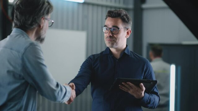 Manager in a suit with a tablet shaking hands with a male client after car diagnostics in a car service, and looking at the camera