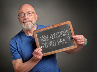 What questions do you have? Senior man, teacher, presenter or mentor, is holding a slate blackboard with white chalk text. Education and communication concept.