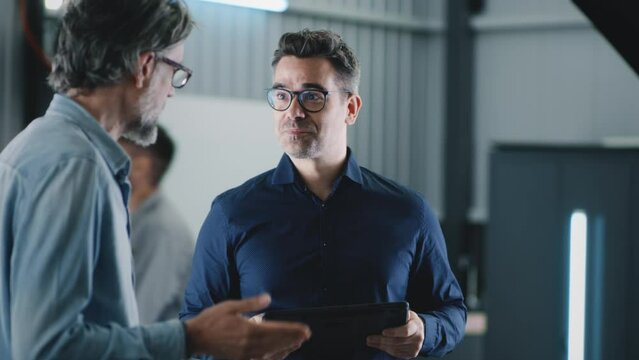 Handheld shot in a repair shop of car service a manager with a tablet greeting a male client talking to him, and consulting about repairing a car after a crash