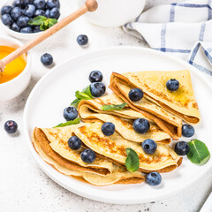Crepes with blueberries and honey in white plate at white table.